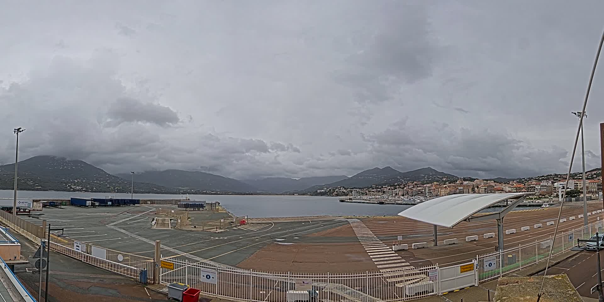 An overcast sky hangs over a harbor with a large empty parking lot, a town in the distance, and mountains beyond.