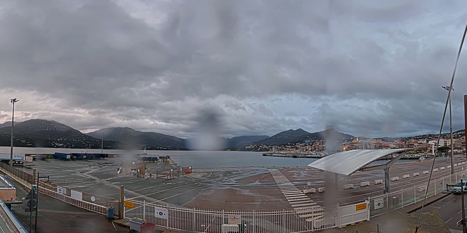 A rain-soaked port area with mountains in the background under an overcast sky.