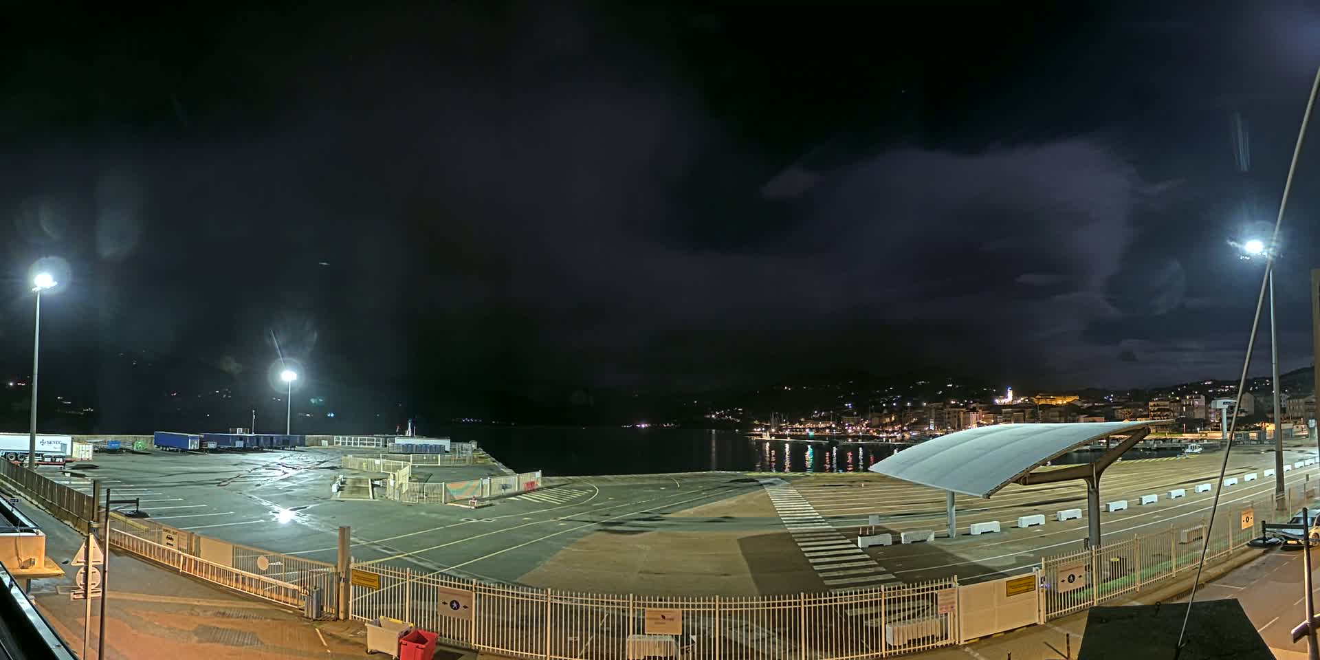 A nighttime view of an empty port area with a few trucks parked, under a dark, cloudy sky.