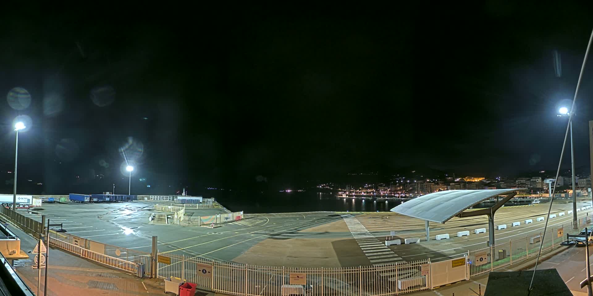 A nighttime view of an empty port area with parked trucks, illuminated by several bright lights, overlooking a dark body of water with a distant town's lights visible.