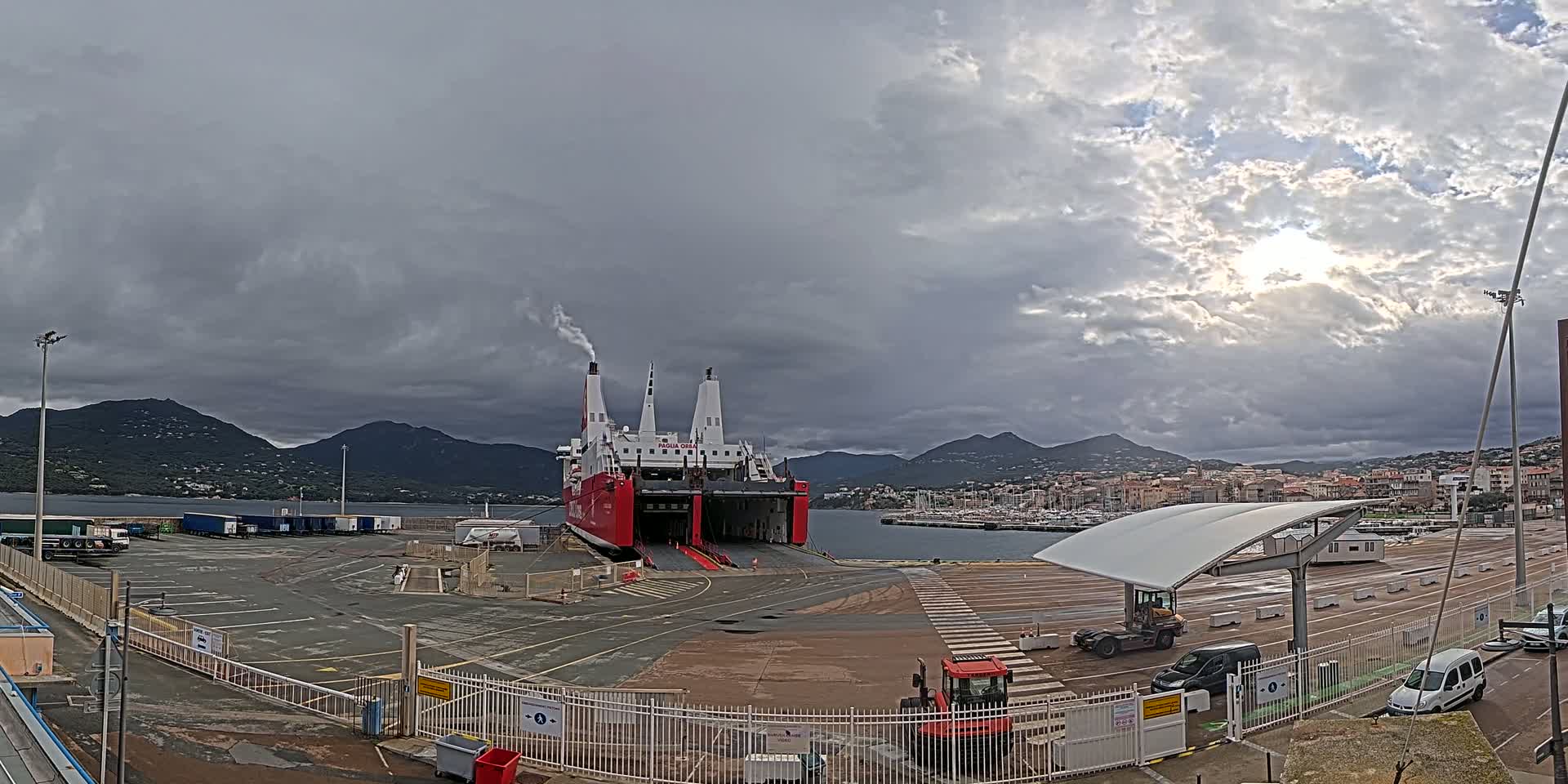 A large red and white ferry is docked at a port under a cloudy sky, with mountains visible in the background.