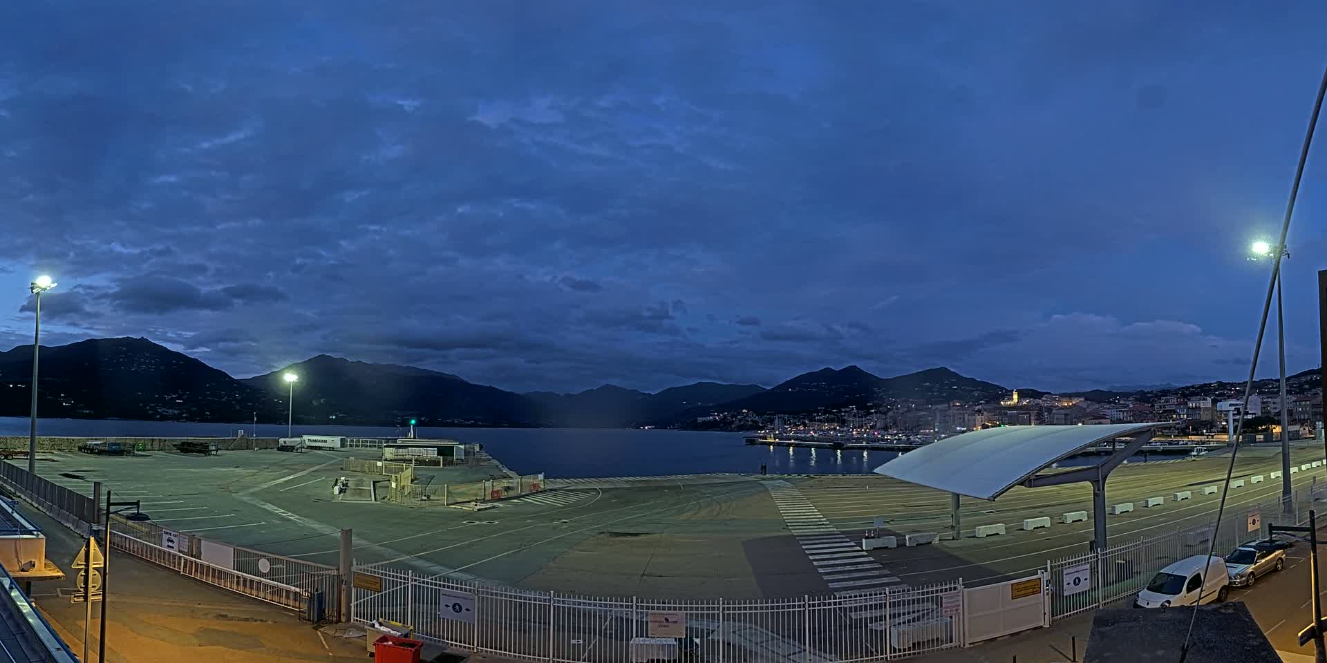 A large, empty paved area at a harbor is bordered by fences, under a twilight sky with scattered clouds, overlooking a calm body of water and distant mountains with a town visible on the other side.