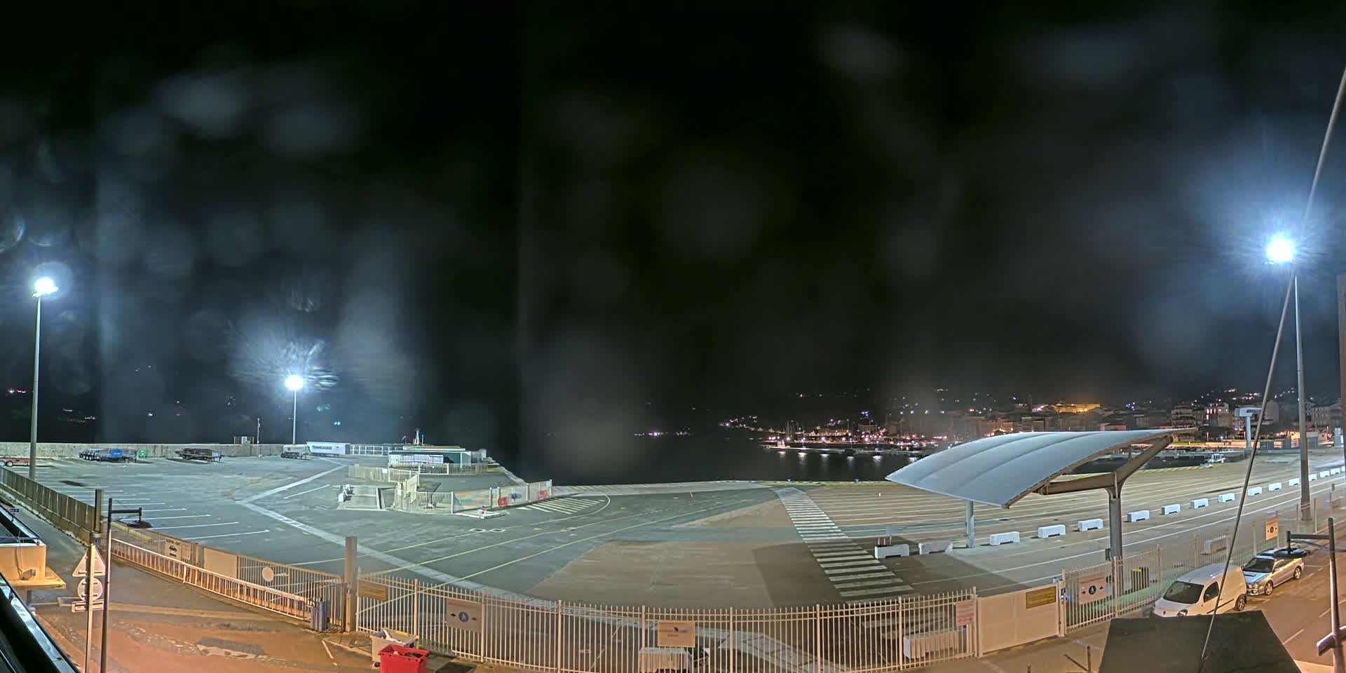 A nighttime view of an empty port area with rain obscuring the view, illuminated by several bright lights, overlooks a dark body of water with a distant town's lights visible.