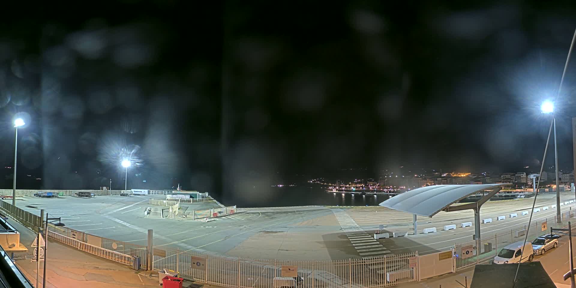 A nighttime view of an empty port area with a few parked vehicles, illuminated by bright lights under a rainy sky.