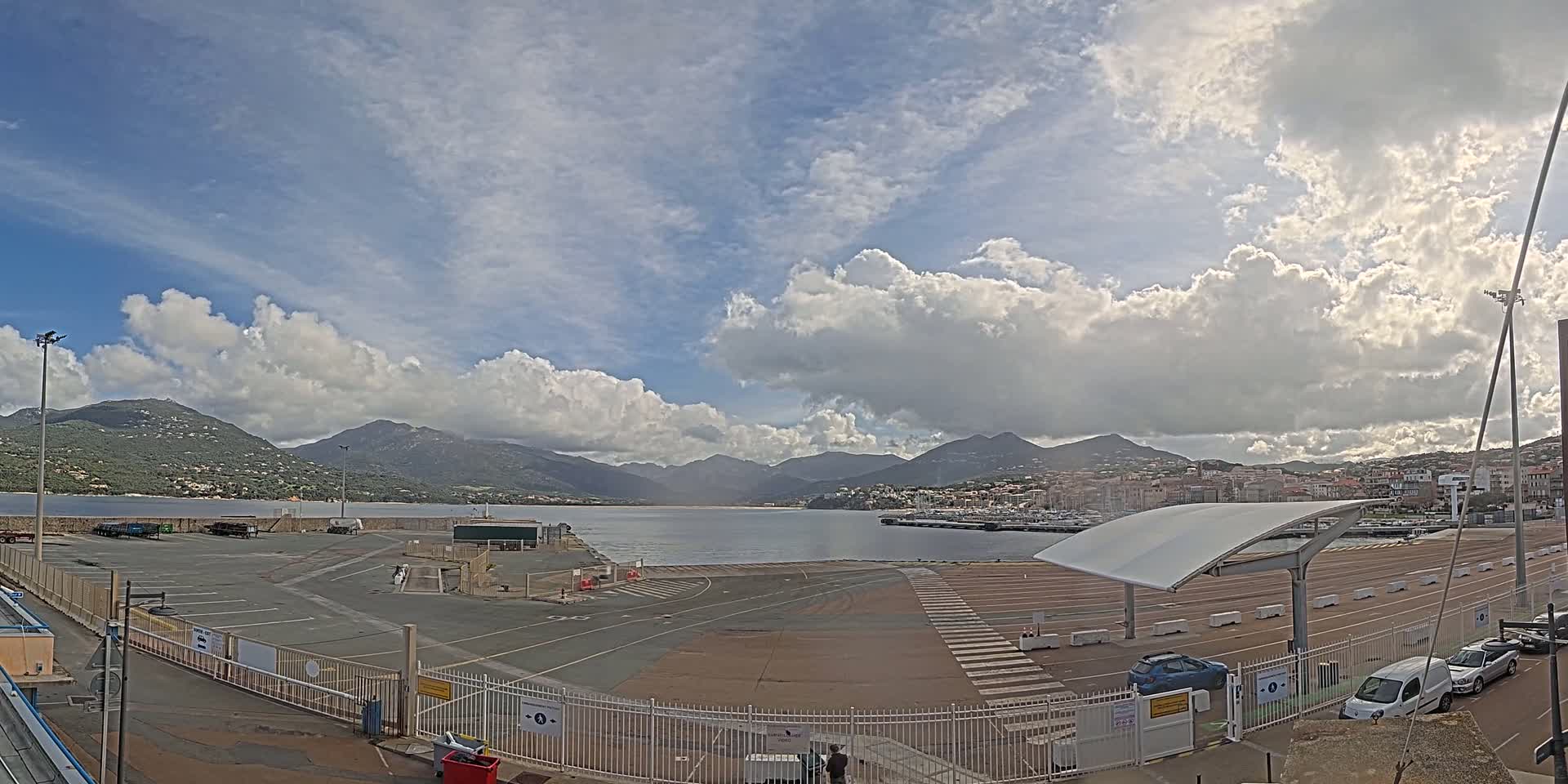 A mostly sunny day with fluffy clouds over a calm body of water, mountains in the background, and an empty parking lot in the foreground near a port.