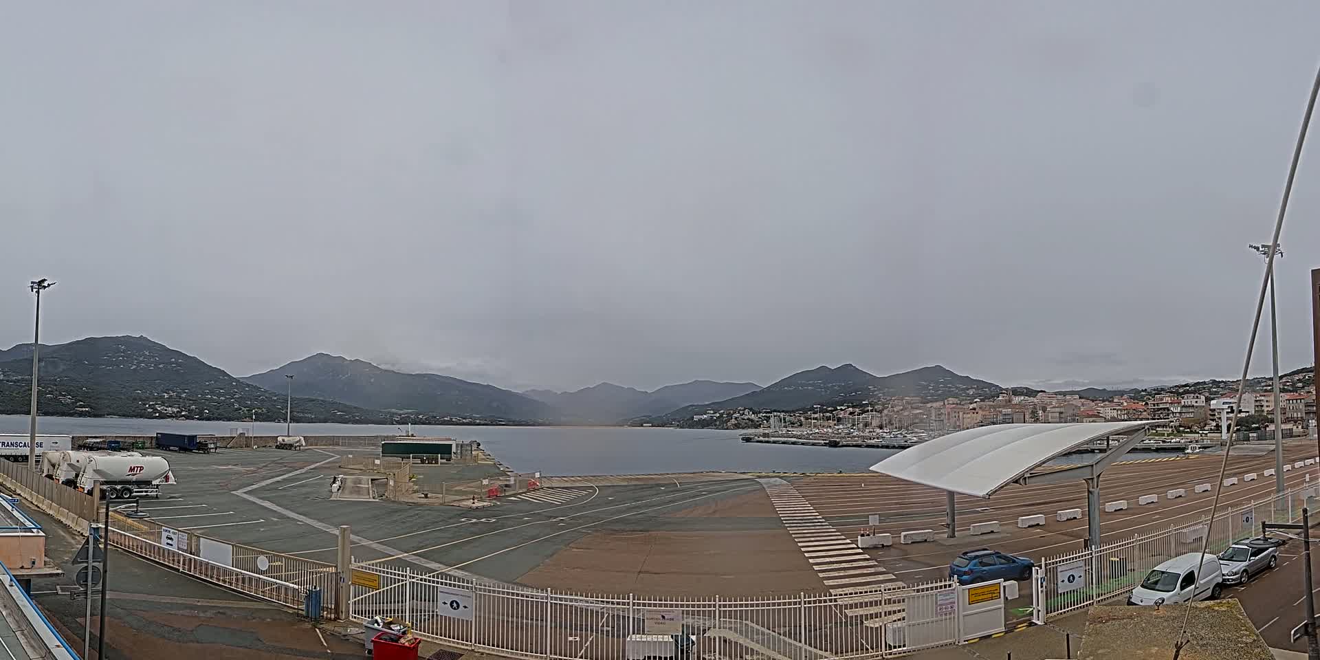An overcast day shows a mostly empty port area with a few vehicles parked near a body of water and mountains in the background.