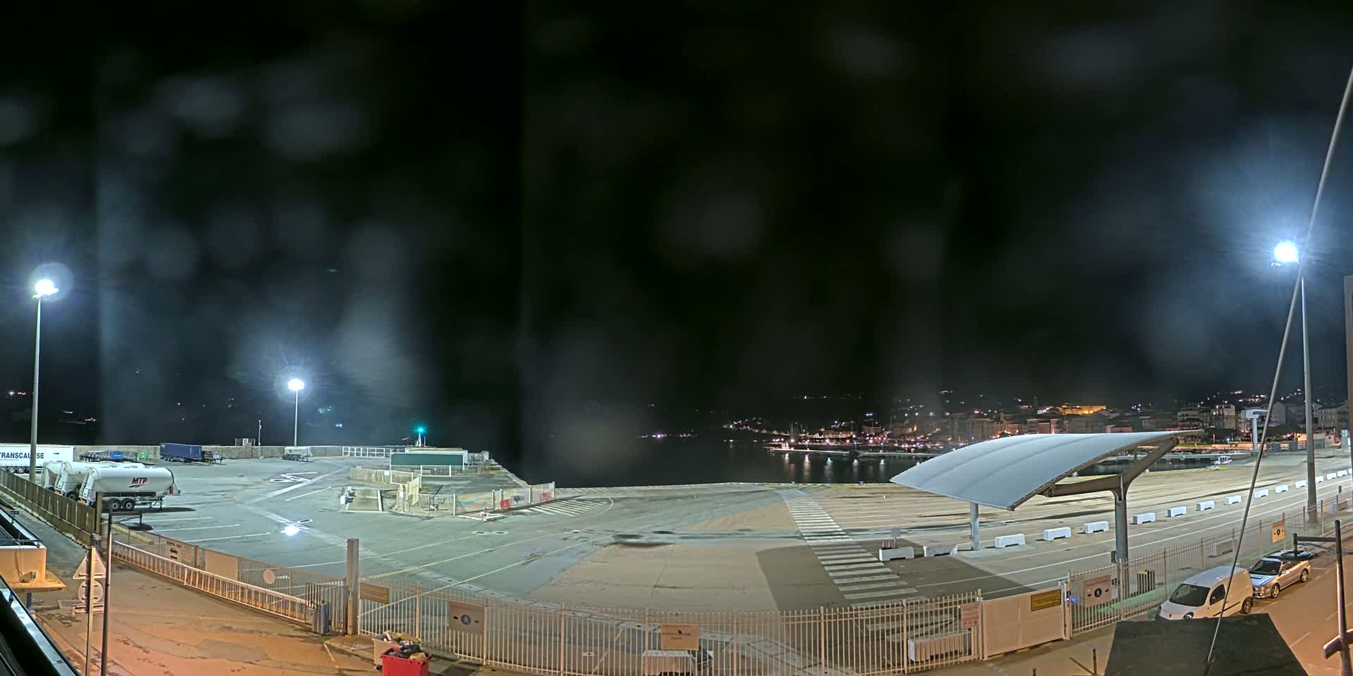 A nighttime view of a mostly empty port area with a few vehicles and some distant lights across the water under hazy conditions.