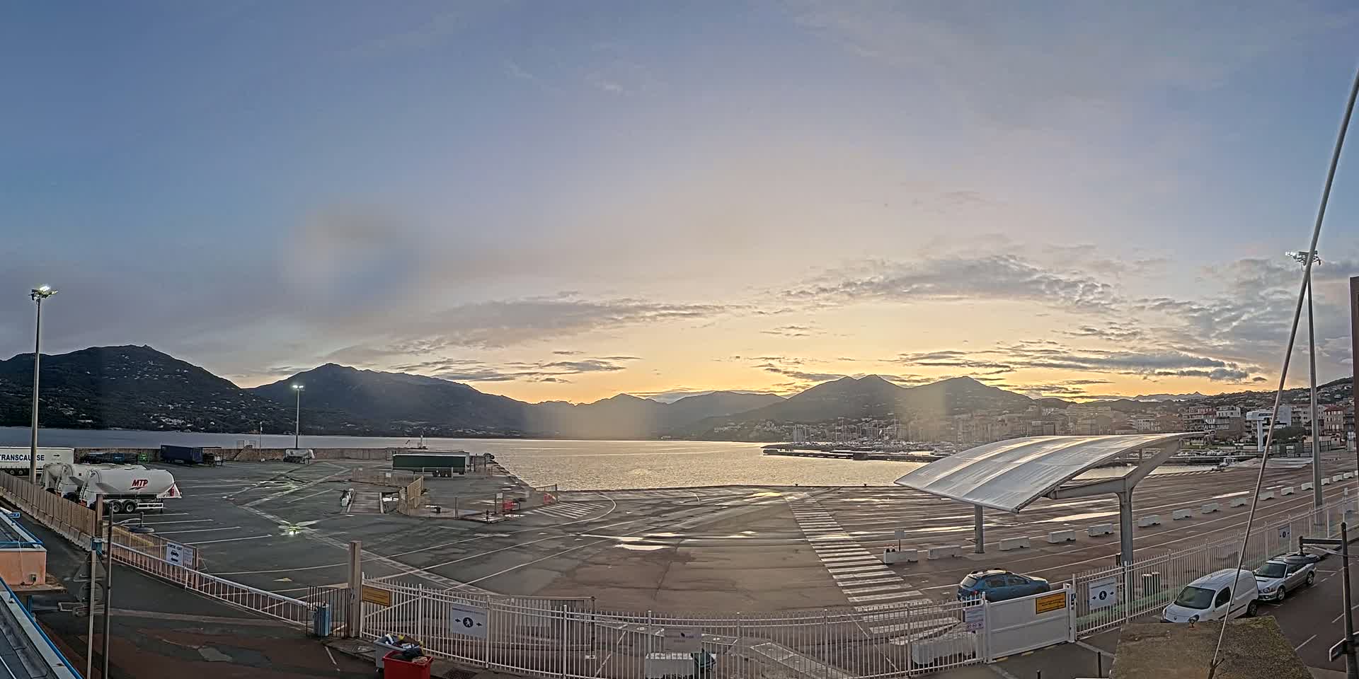A mostly empty port parking lot with a few cars and trucks, overlooked by mountains at sunrise under a partly cloudy sky.