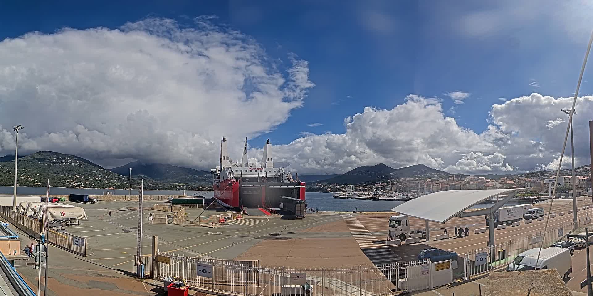 A large red and white ferry is docked at a port under a partly cloudy sky, with mountains and a town visible in the distance.