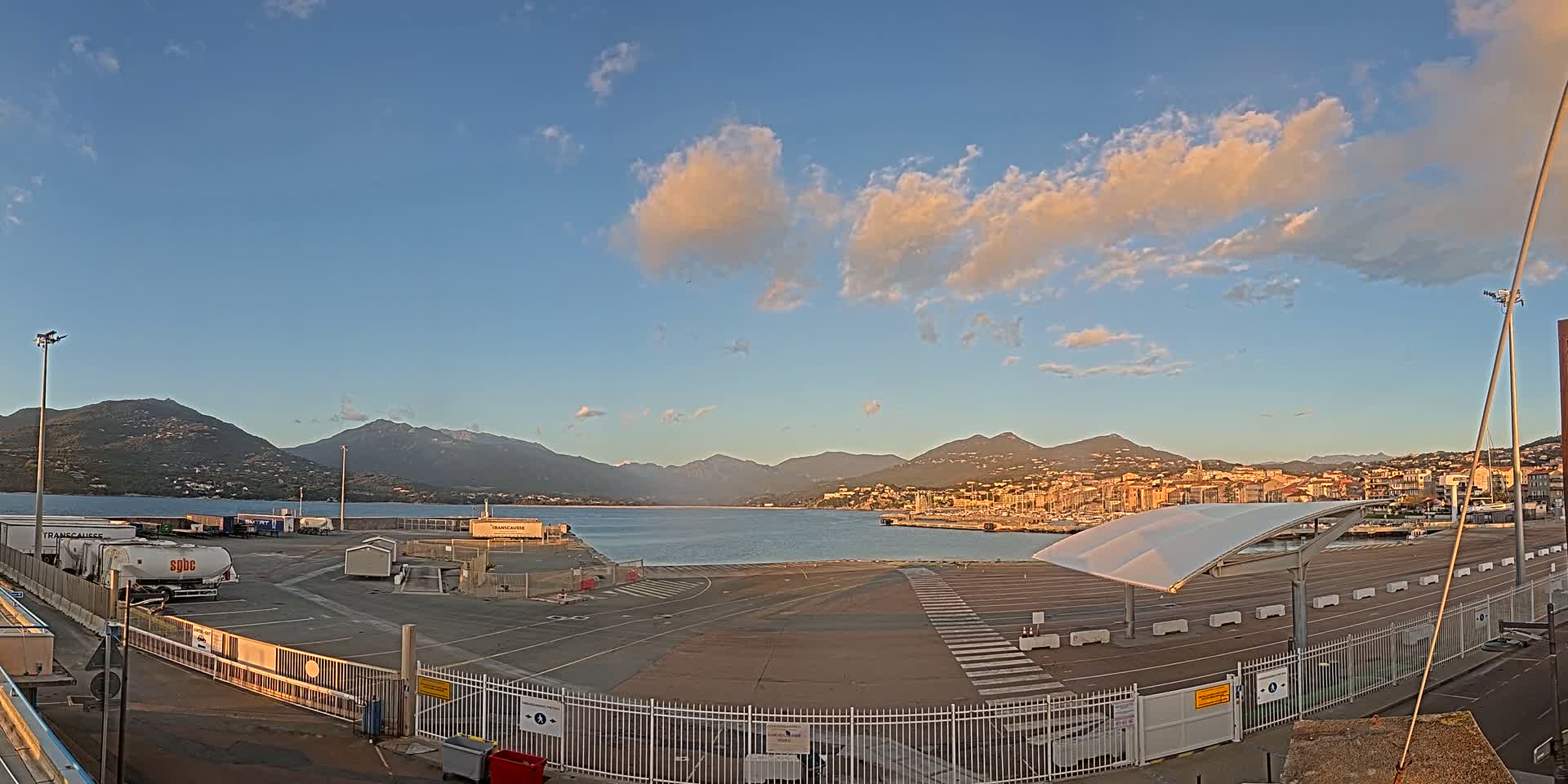 A mostly empty port area with a few trucks and a covered waiting area, overlooks a calm body of water and mountains under a partly cloudy, sunset sky.