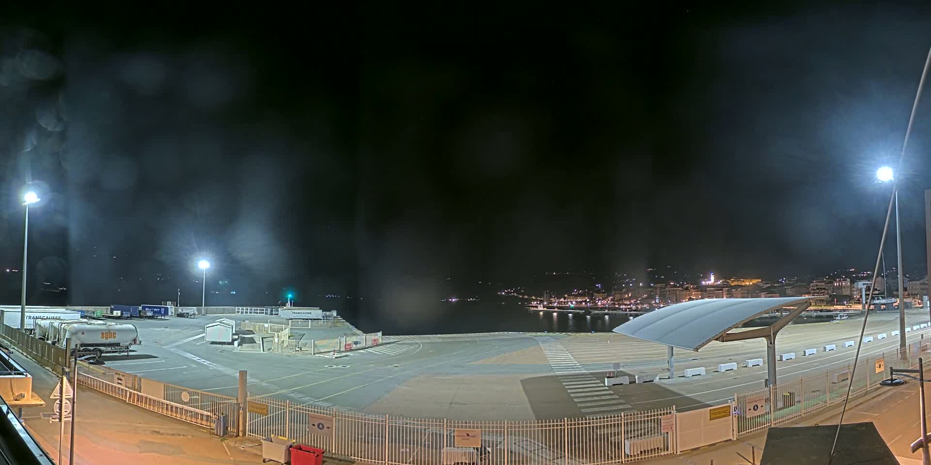 A nighttime view of an empty port area with several trucks parked, lit by bright lights, overlooking a calm body of water and a distant city skyline.