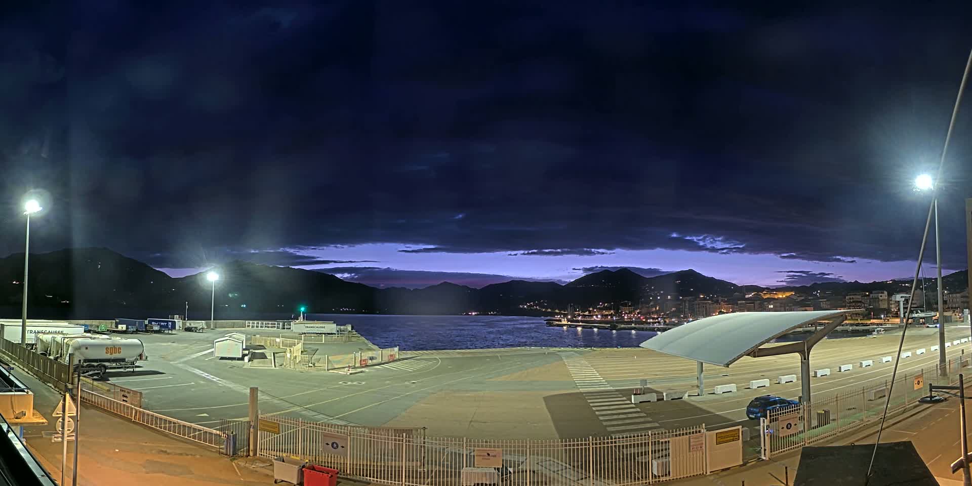 An empty, well-lit port area at twilight, under a dark, cloudy sky, overlooks a calm body of water and distant mountains.