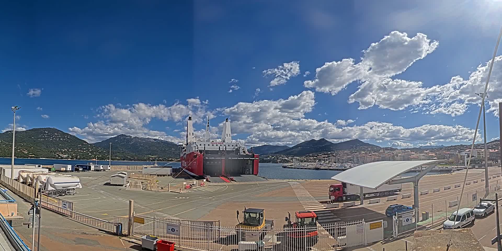 A large red and white ferry is docked at a port under a partly cloudy blue sky, with mountains in the background and various vehicles and equipment in the foreground.