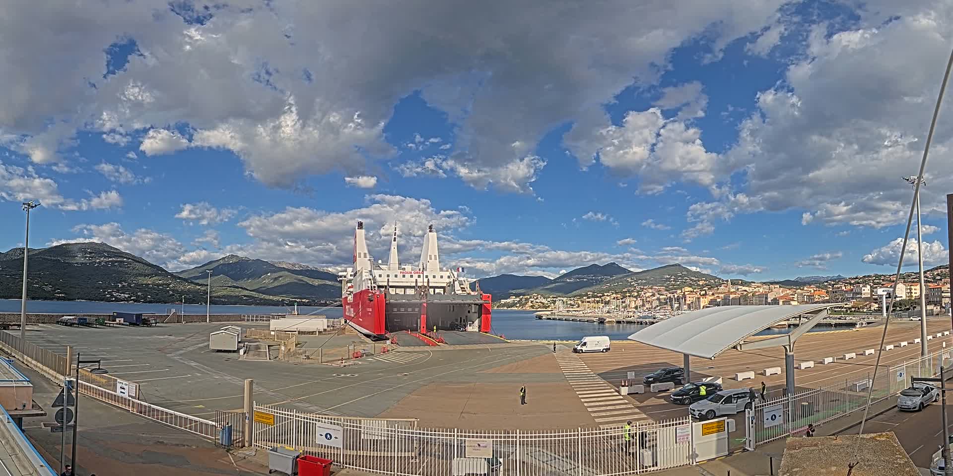 A large red and white ferry is docked at a port under a partly cloudy sky, with mountains and a town visible in the background.