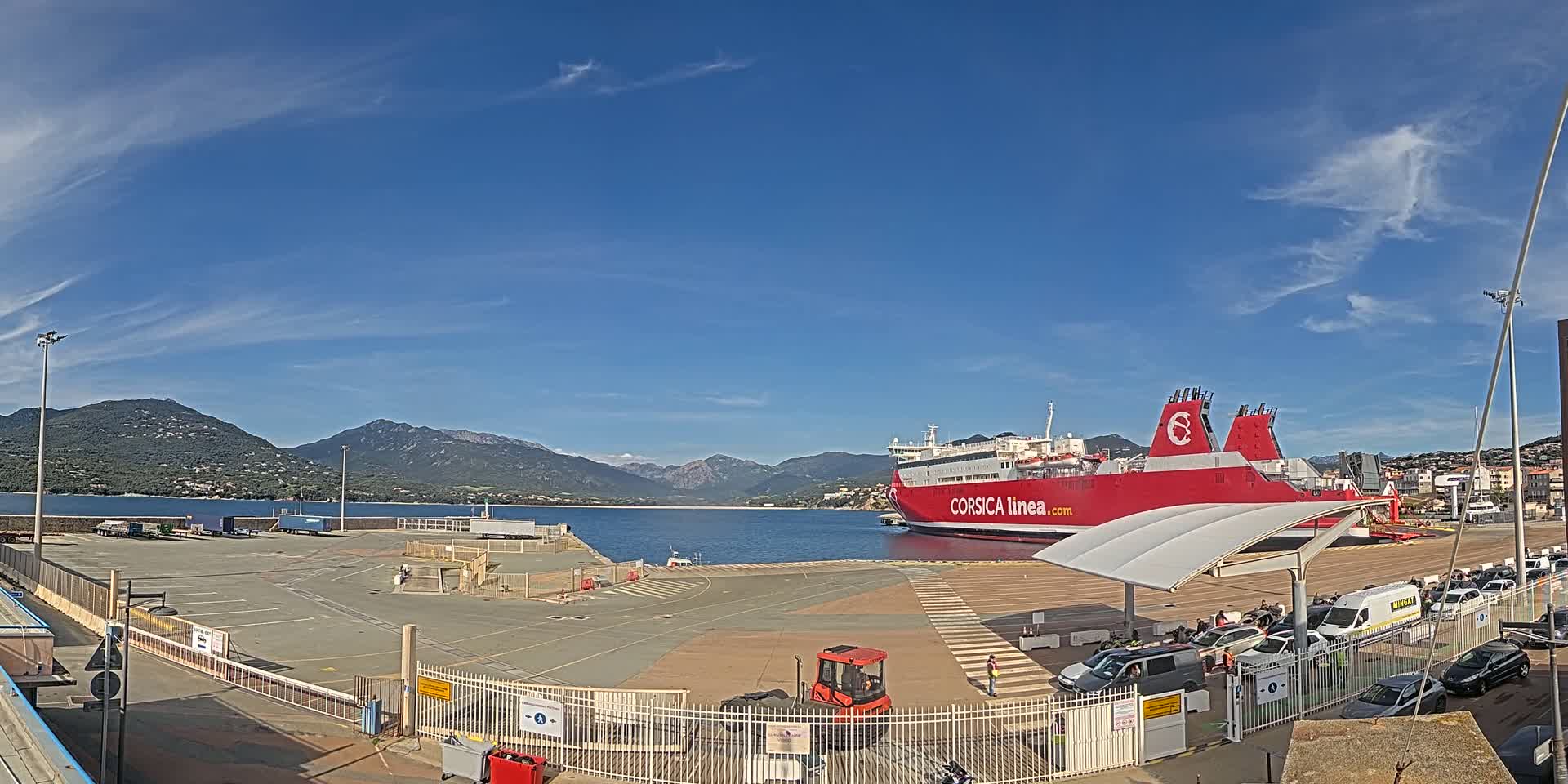 A large red ferry is docked at a port under a clear blue sky, with mountains visible in the distance.