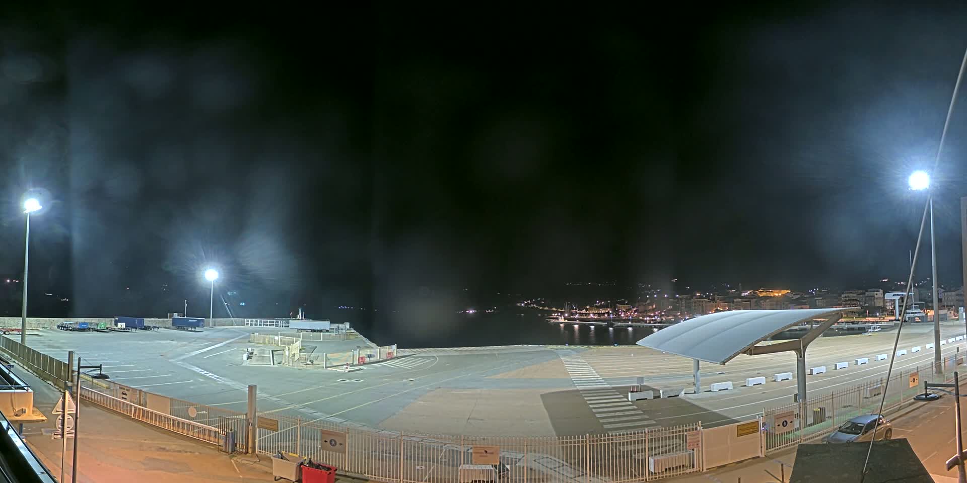 A nighttime view of an empty port area with hazy conditions, showing a paved lot, some parked vehicles, and a distant town illuminated across the water.