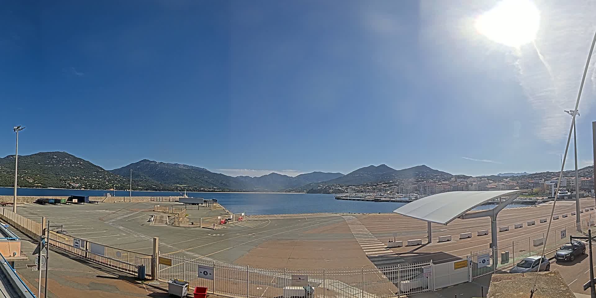 A sunny day reveals a harbor with a large, empty paved area, bordered by a fence, with mountains and a town in the background.