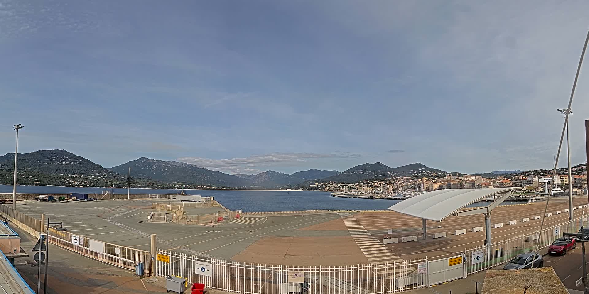 An empty port area with a few cars parked near a gated fence, overlooking a calm body of water and mountains under a mostly clear sky.