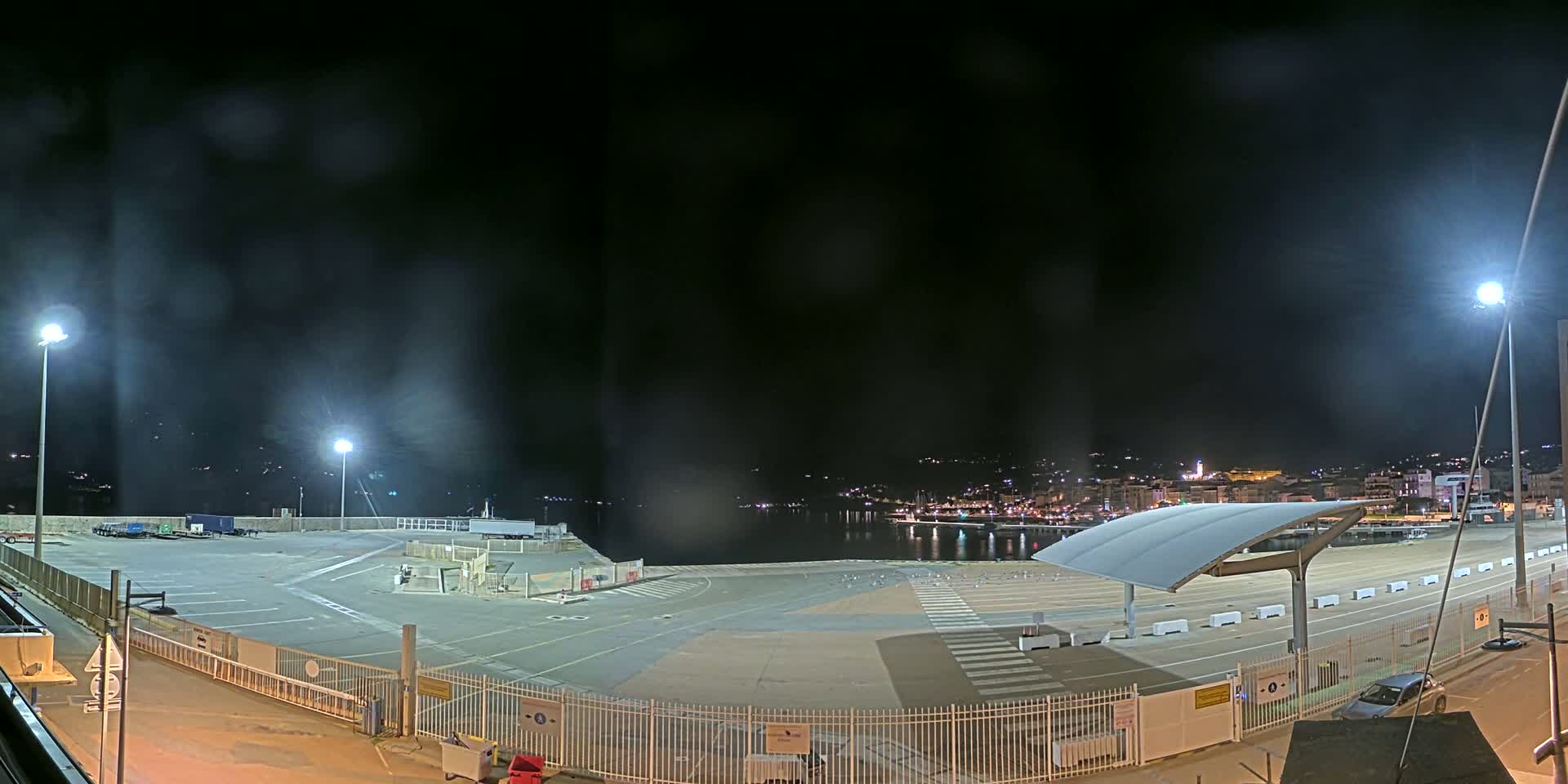 An empty, brightly lit port area at night overlooks a dark, calm body of water and a city skyline in the distance.