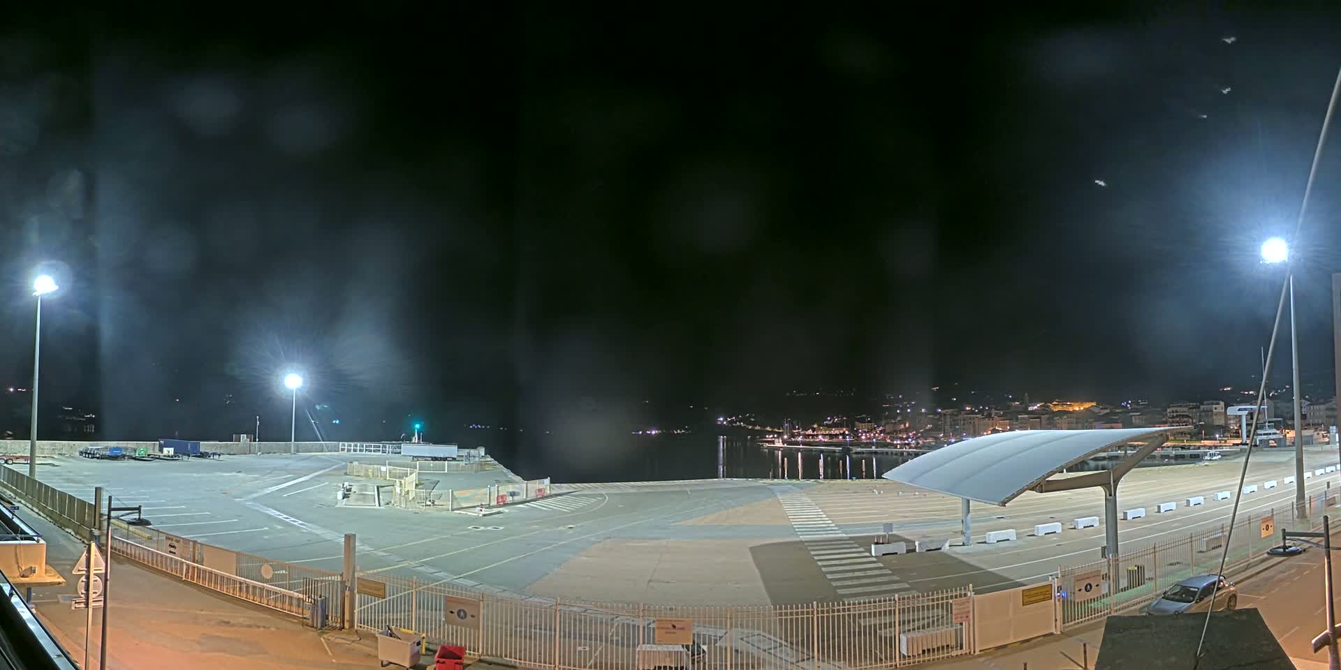 An empty, brightly lit nighttime port area with a partially covered waiting area and a distant, faintly visible town across the water under a hazy sky.