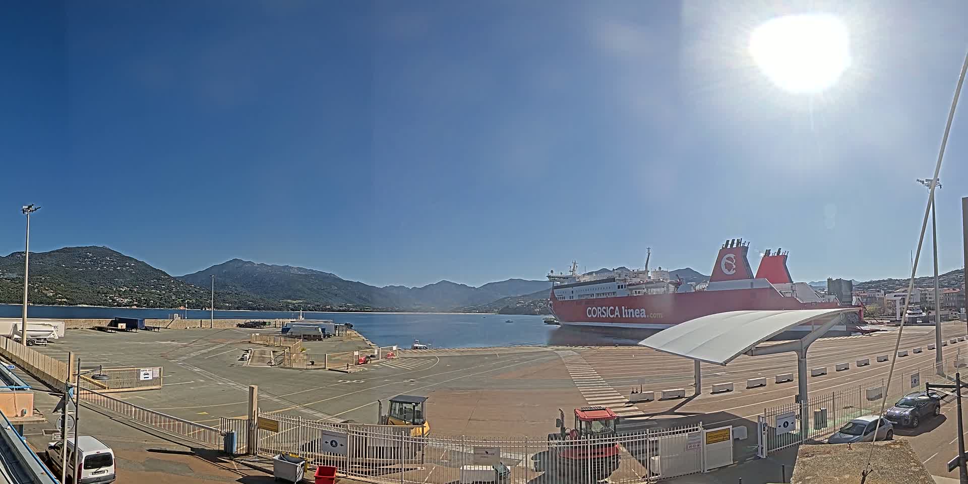 A large red ferry is docked at a port on a sunny day, with mountains visible in the background.