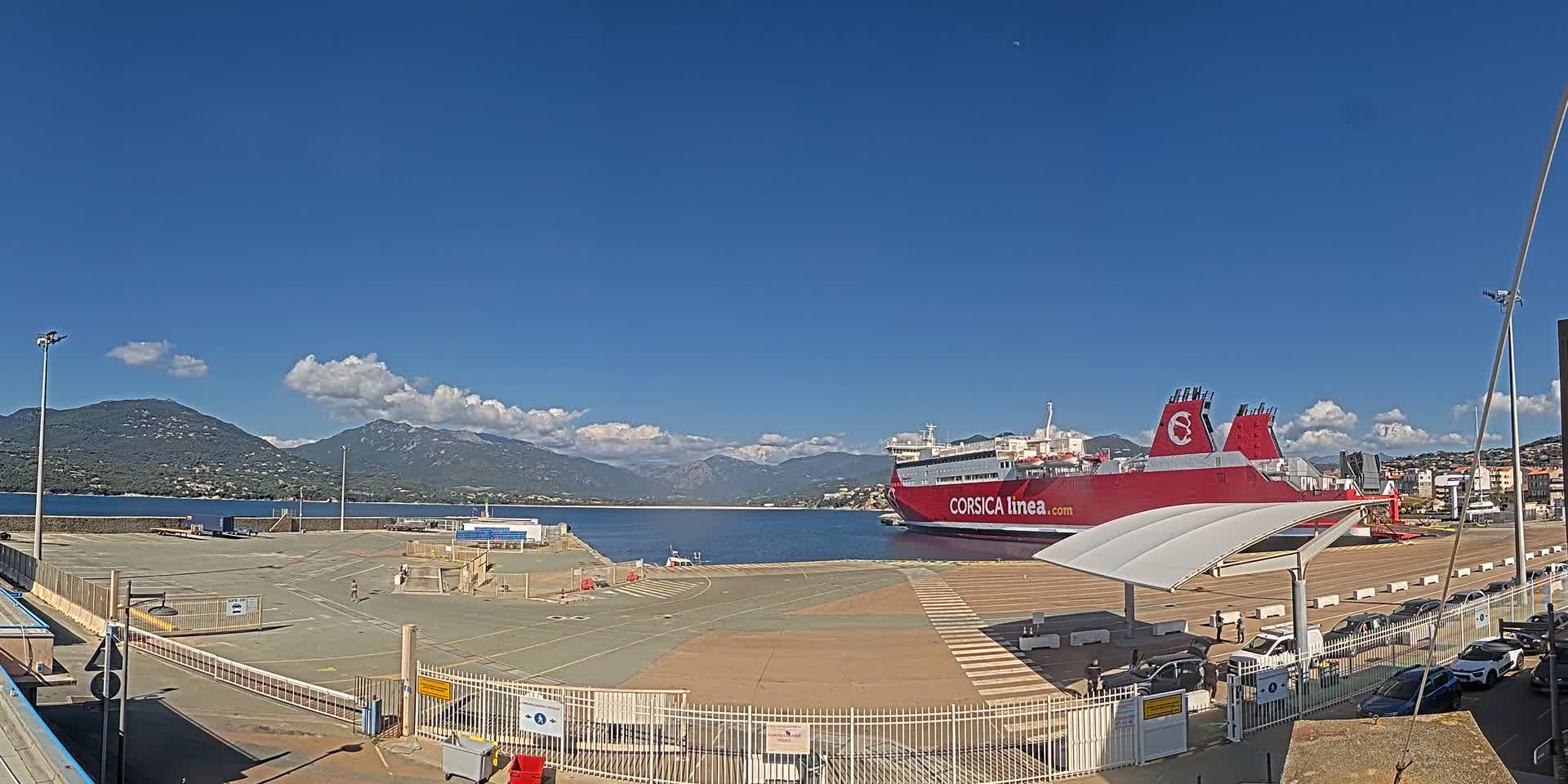 A large red ferry is docked at a port, under a mostly sunny sky with some clouds, mountains are visible in the background.