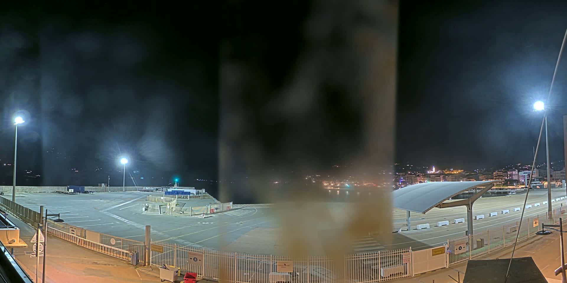 A nighttime view of a mostly empty port area with hazy atmospheric conditions, illuminated by several bright lights, and a city skyline visible in the distance.