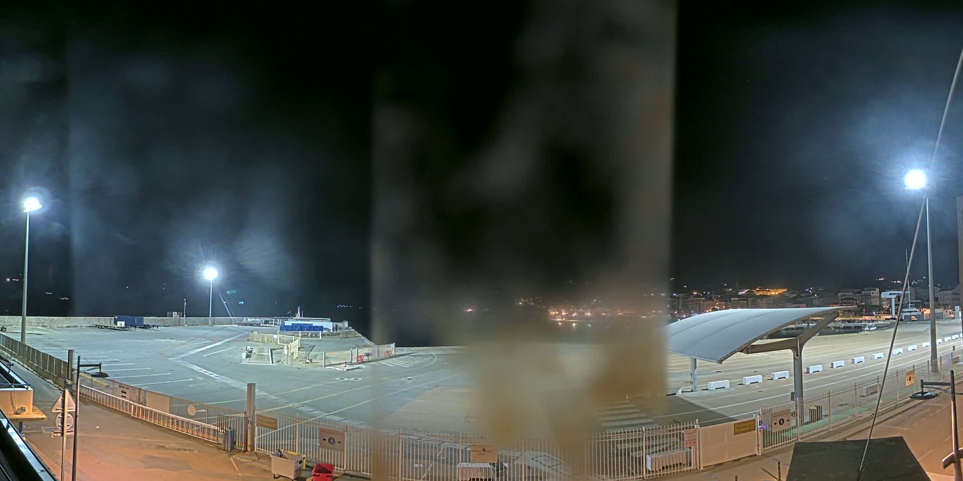 A nighttime view of an empty port area with hazy or foggy conditions, illuminated by bright lights, and a town visible in the distance.