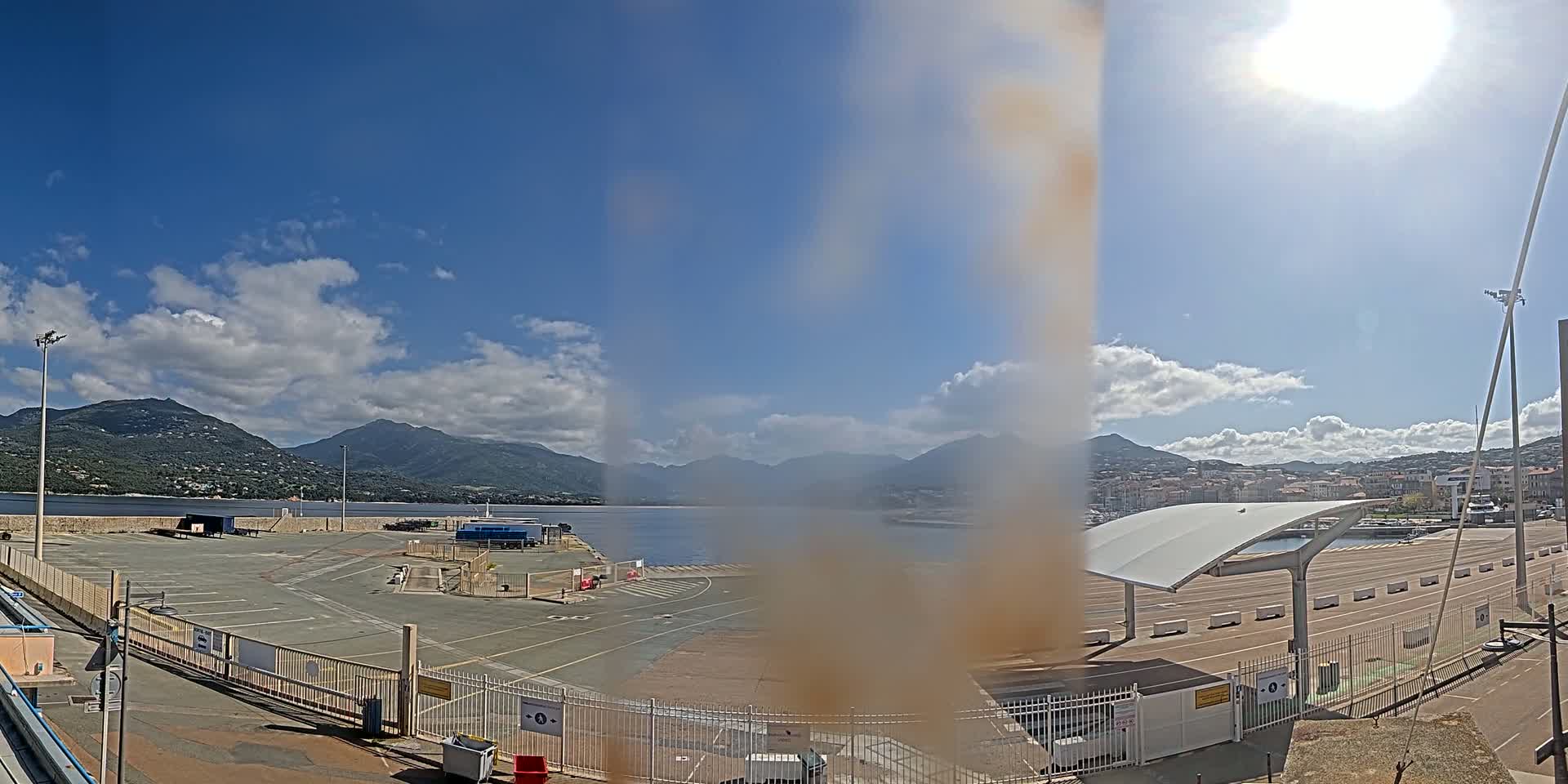 A sunny day with partly cloudy skies shows a harbor's empty parking lot, a waterfront, and mountains in the distance.