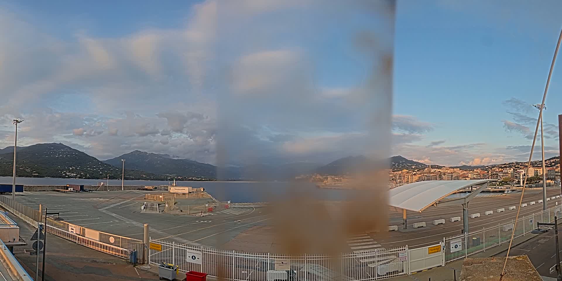 A partly cloudy sky overlooks a port area with a calm body of water, mountains in the background, and a mostly empty parking lot.