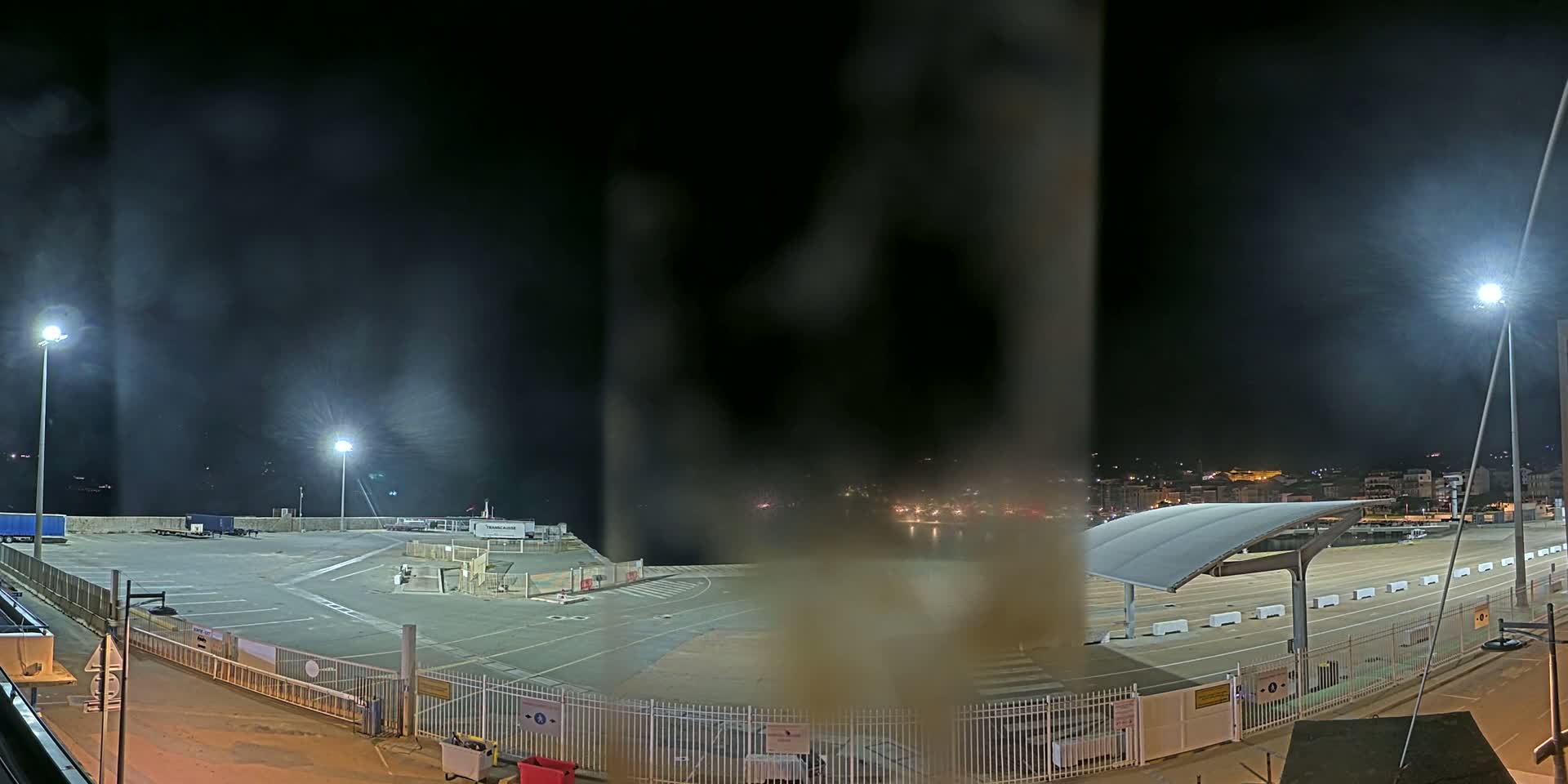 A nighttime view of an empty port area with hazy or foggy conditions, illuminated by several bright lights, overlooking a city in the distance.