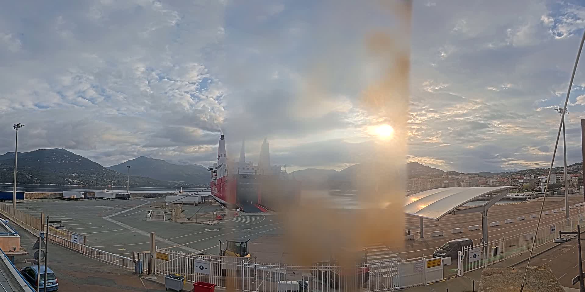A large ferry is docked at a port, under a partly cloudy sky with the sun shining through, and mountains are visible in the distance.