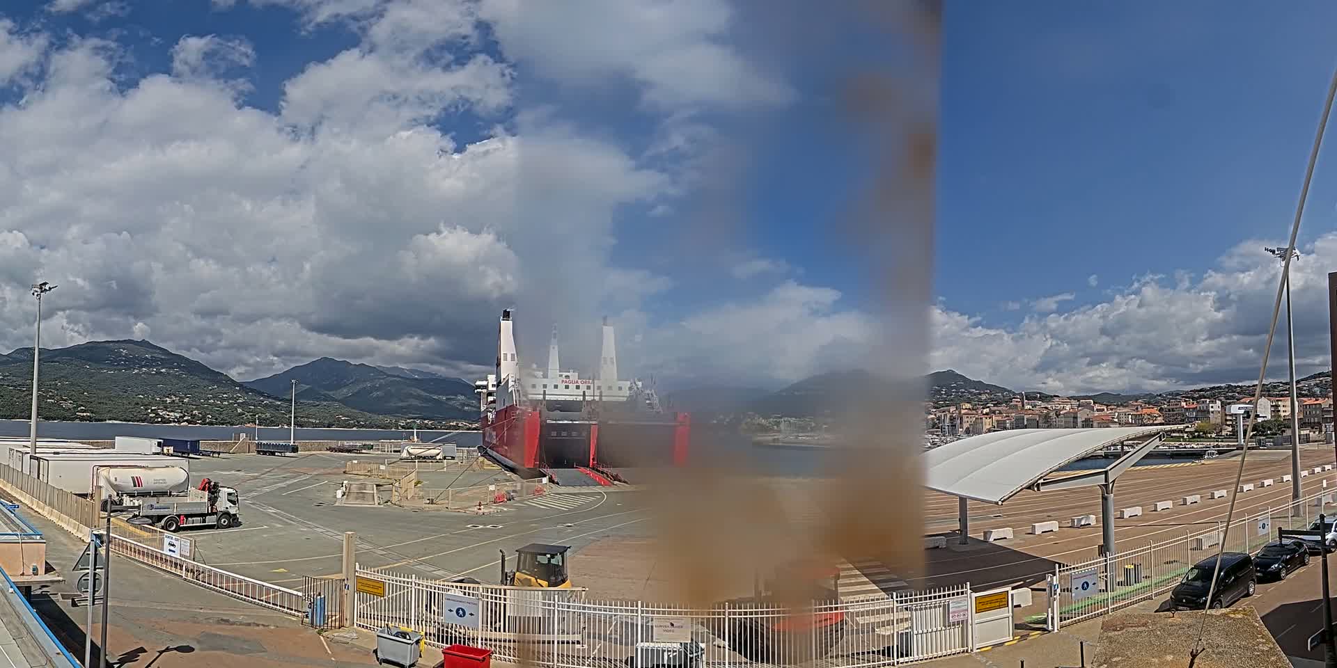 A large red and white ferry is docked at a port under a partly cloudy sky, with mountains visible in the background.
