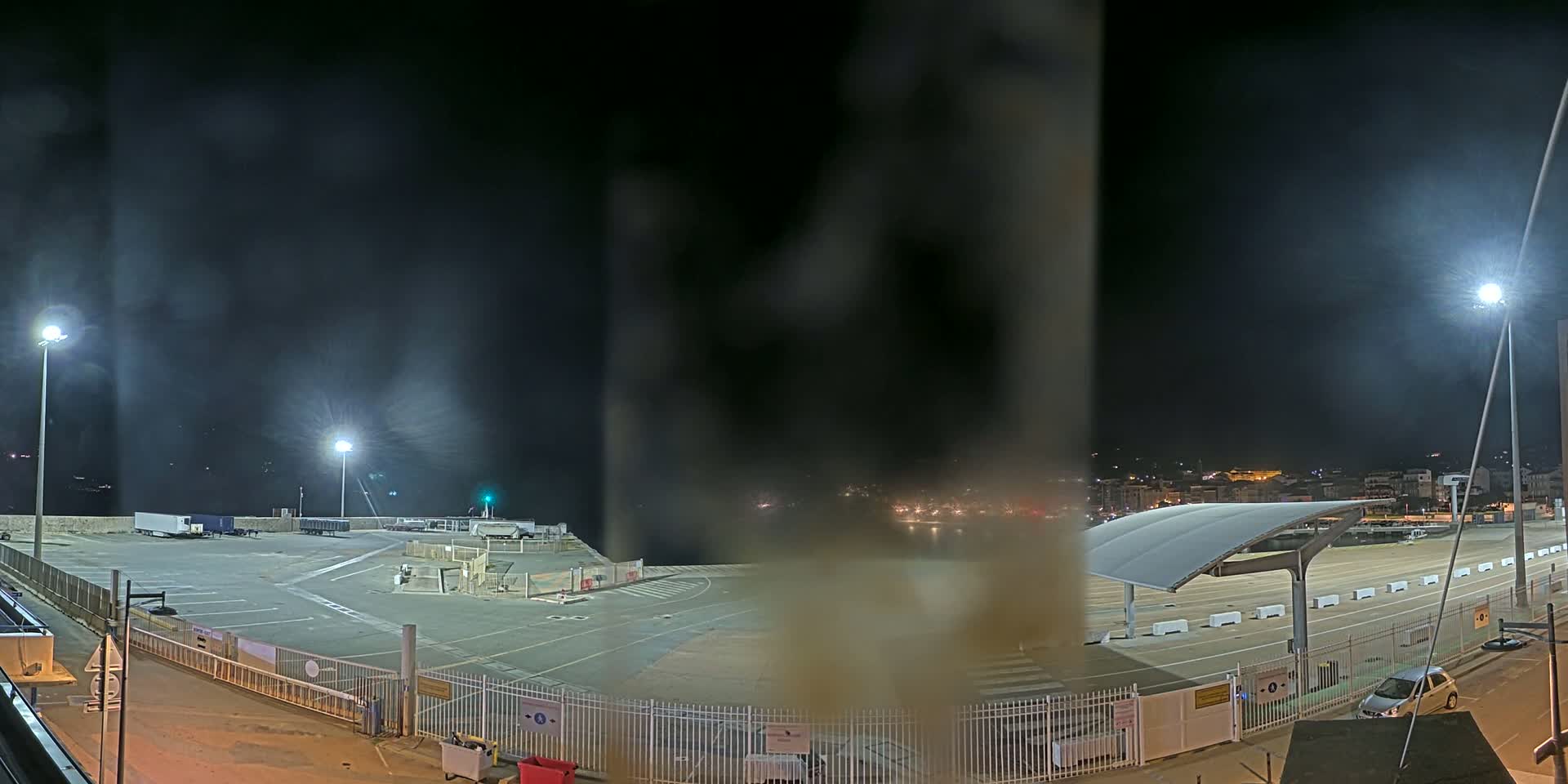 A nighttime view of a mostly empty port area with hazy conditions, illuminated by several bright lights, featuring parked trucks and a few cars.