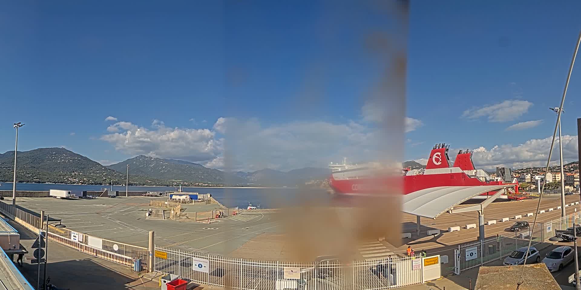 A large red and white ferry is docked at a port, with a partly cloudy blue sky and mountains in the background.