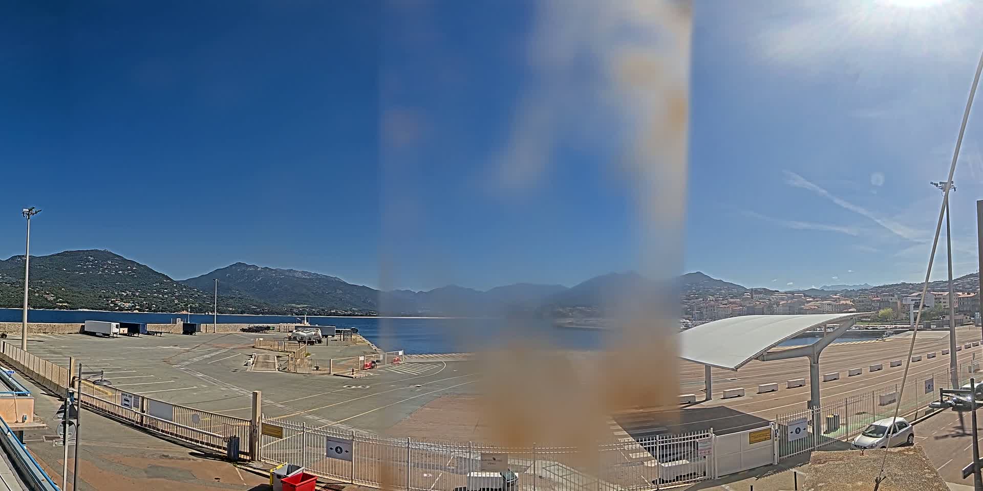 A sunny day reveals a harbor scene with a large empty parking lot, a covered waiting area, and mountains in the background.