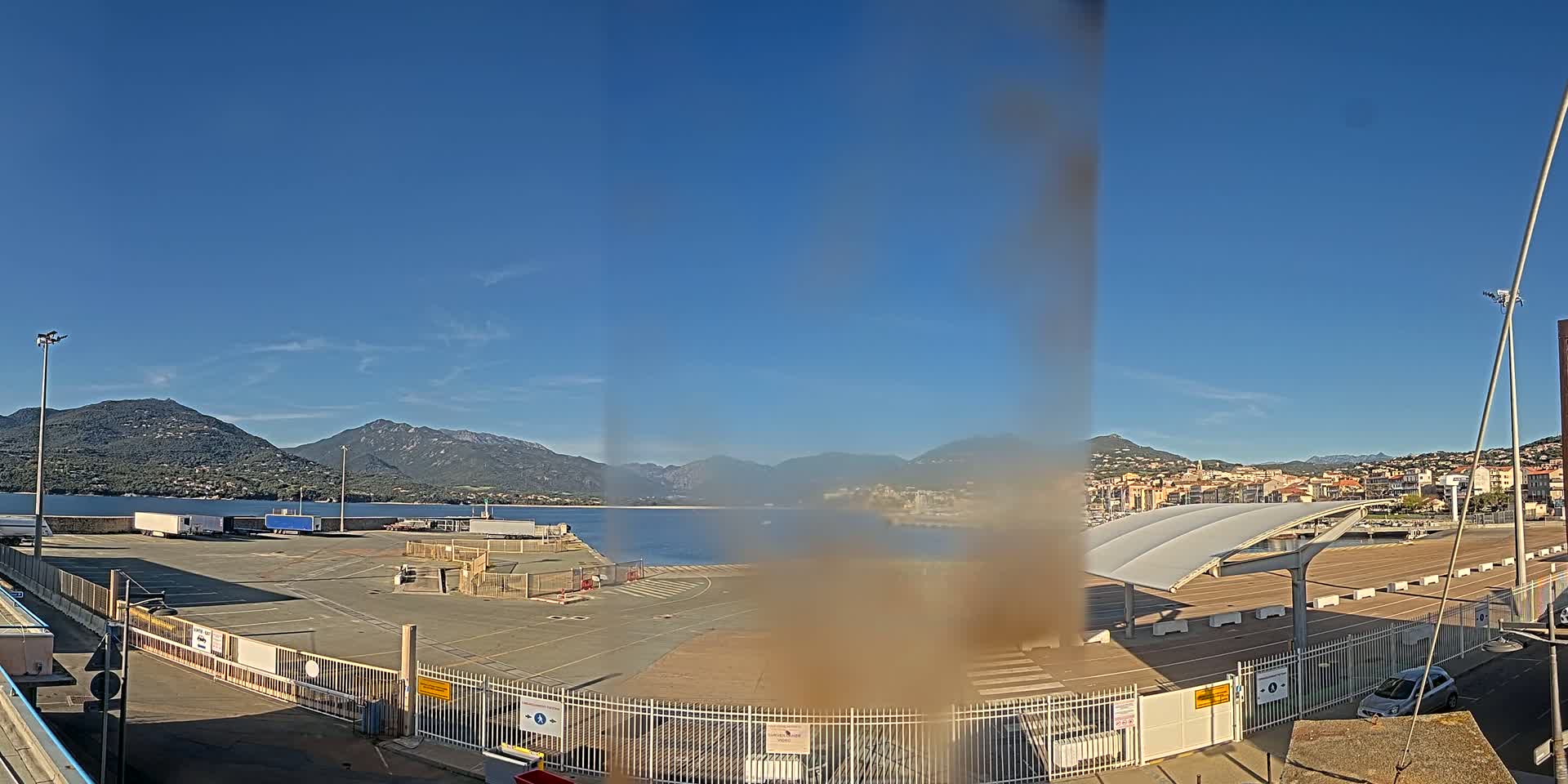 A port area with semi-trucks and a covered waiting area, overlooked by mountains under a clear blue sky.