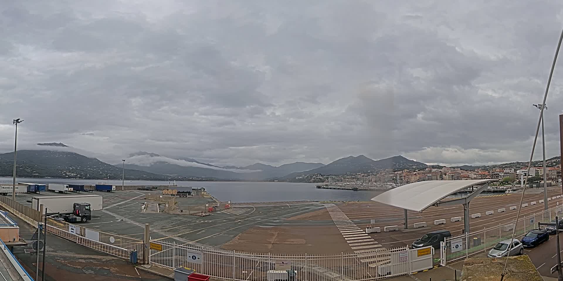 A mostly empty port area with several trucks and cars, under a cloudy sky, overlooks a calm body of water and distant mountains.