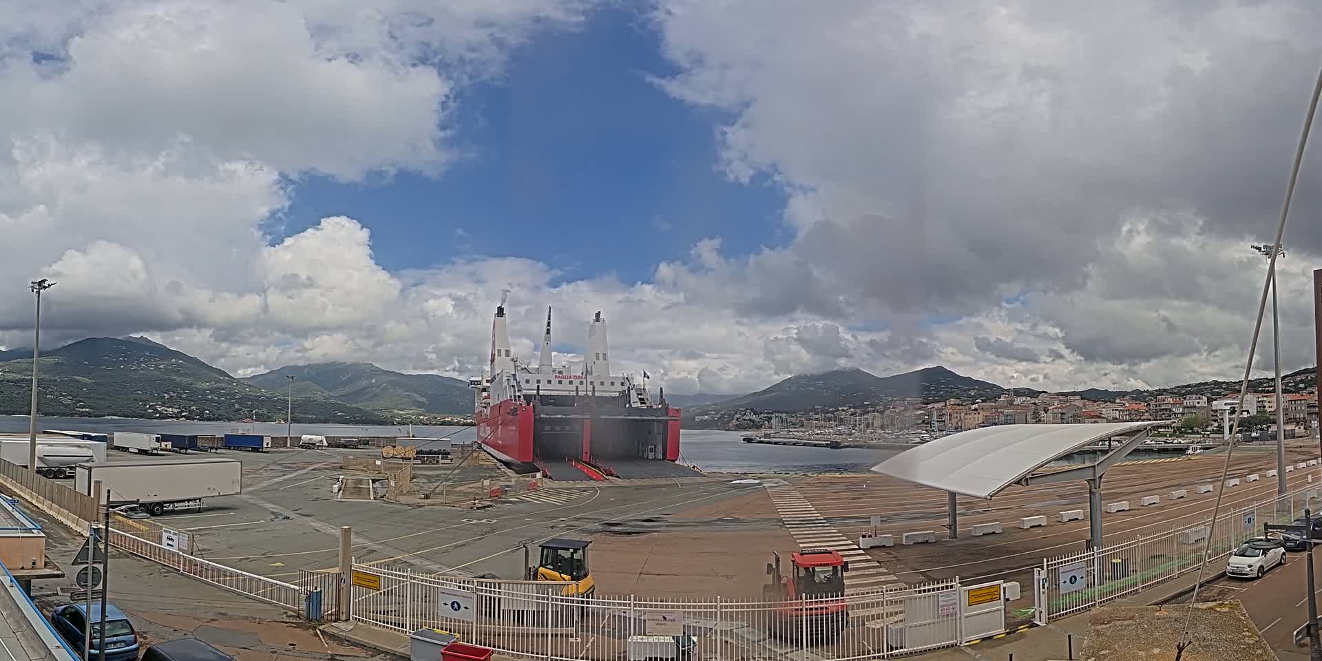 A large red ferry is docked at a port under a partly cloudy sky, with mountains in the background and several vehicles parked nearby.