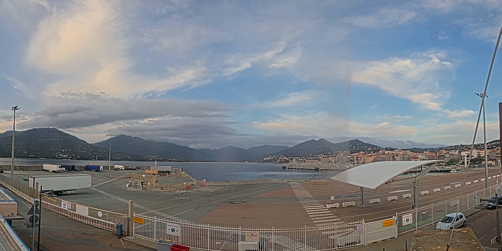 A partly cloudy sky overlooks a port area with several trucks, cars, and a marina, all situated before a range of mountains in the distance.