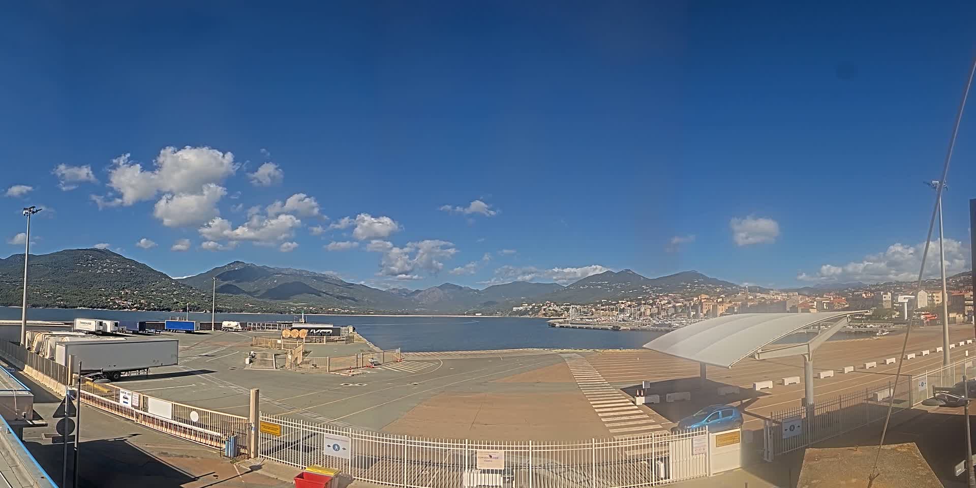 A port area with several trucks and a few cars, under a mostly sunny sky with some clouds, features mountains in the background.