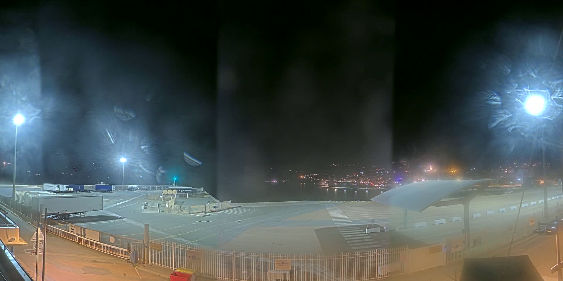 A nighttime view of a mostly empty port area with several semi-trailer trucks, under hazy or foggy conditions.