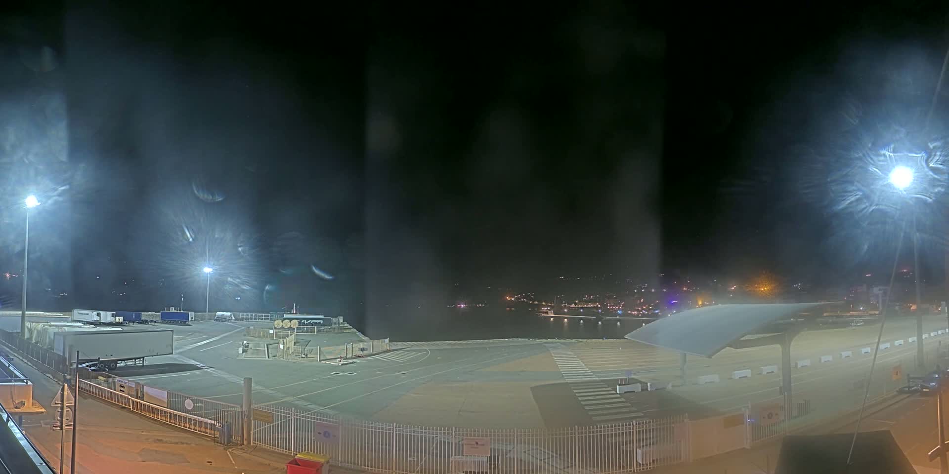 A nighttime view of an empty port area with several parked trucks under bright lights, a body of water visible in the distance, and a slight haze or fog in the air.