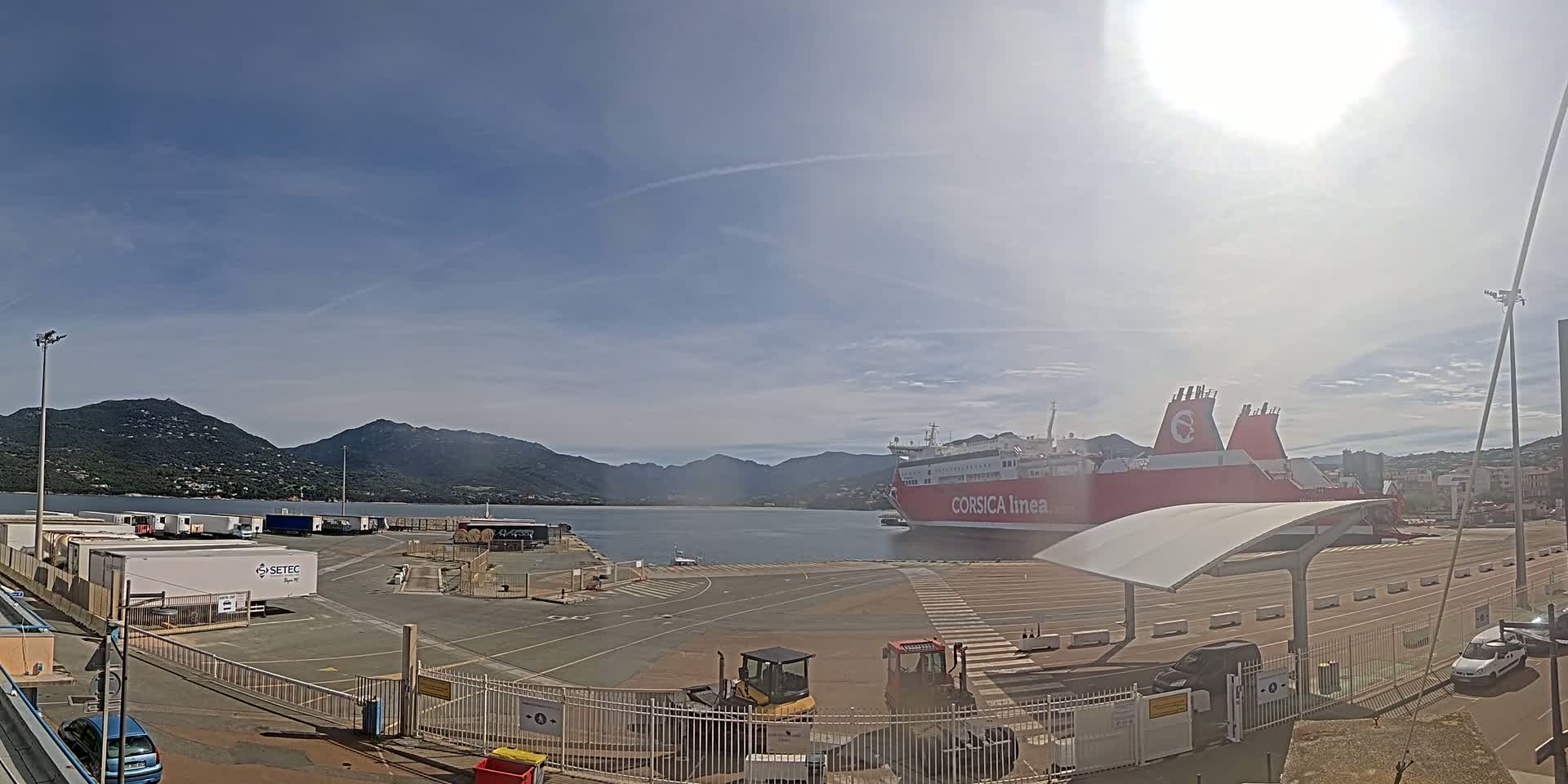 A large red and white ferry is docked at a port, with mountains in the background under a mostly sunny sky.
