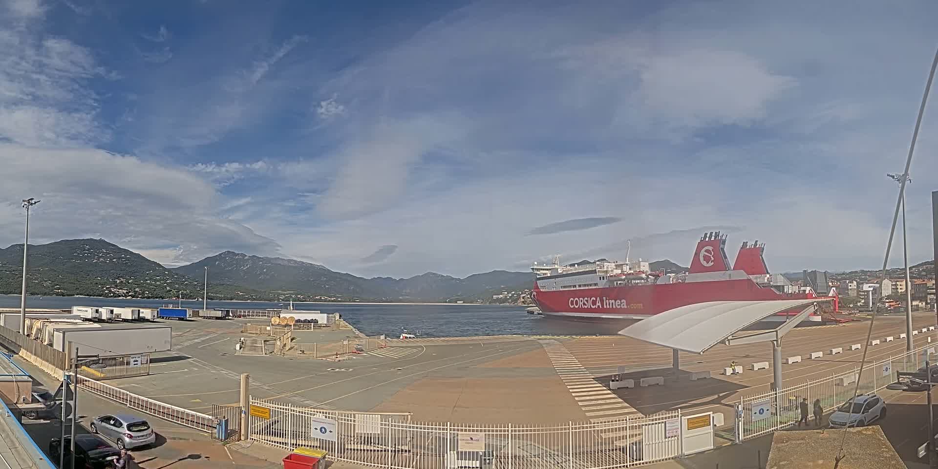 A large red ferry is docked at a port under a mostly sunny sky, with mountains visible in the background.