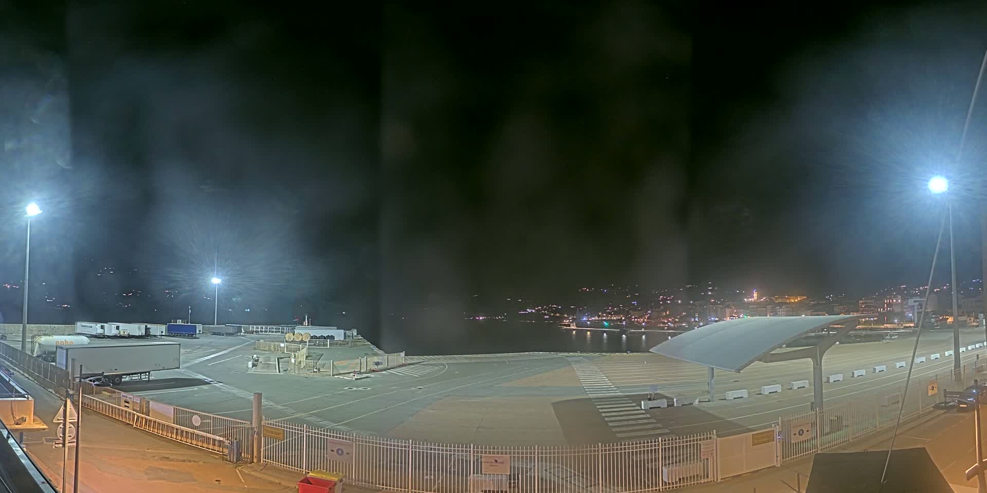 A nighttime view of a mostly empty port area with several trucks, under hazy, dimly lit conditions, overlooking a dark body of water and a distant town.