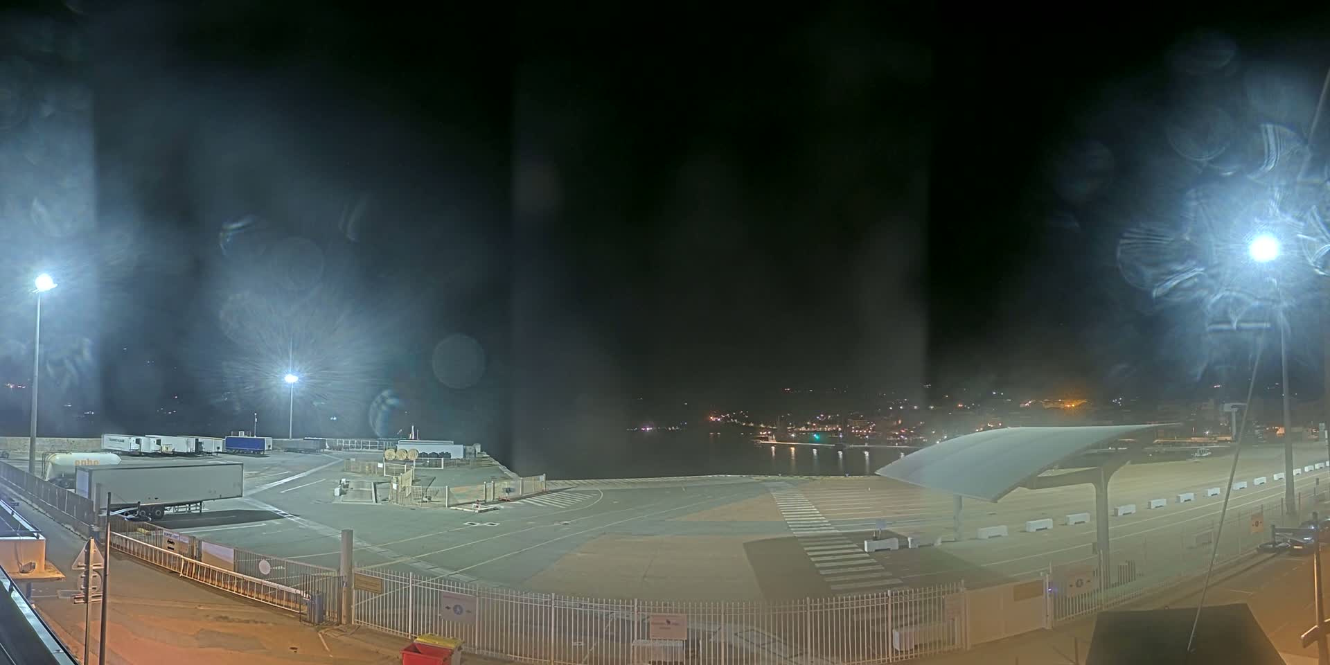 An empty, brightly lit nighttime port area with several trucks parked, a body of water in the background, and light rain or mist obscuring the view.