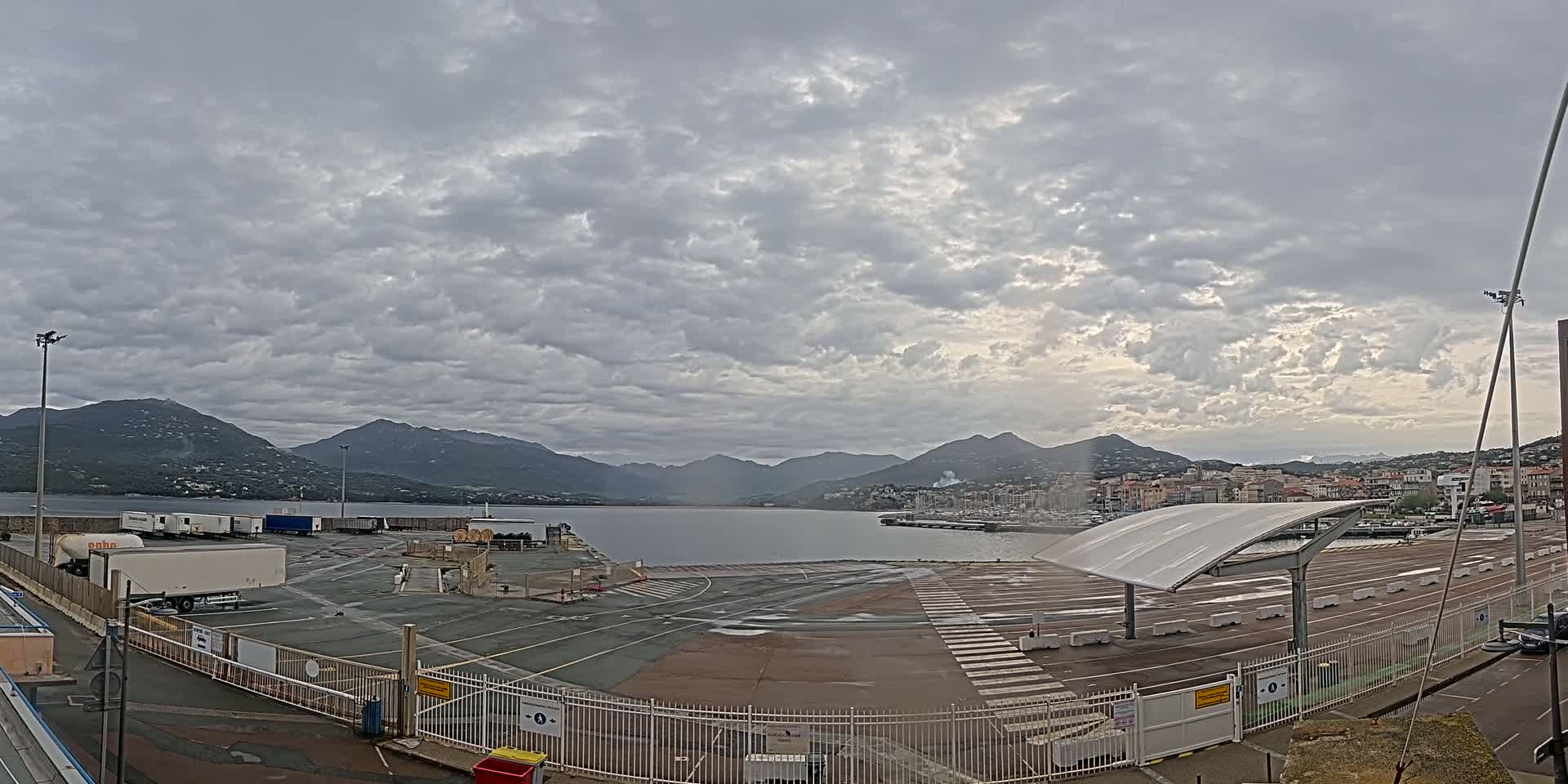 A mostly empty port area with several large trucks, under a cloudy sky, overlooks a calm body of water and distant mountains.
