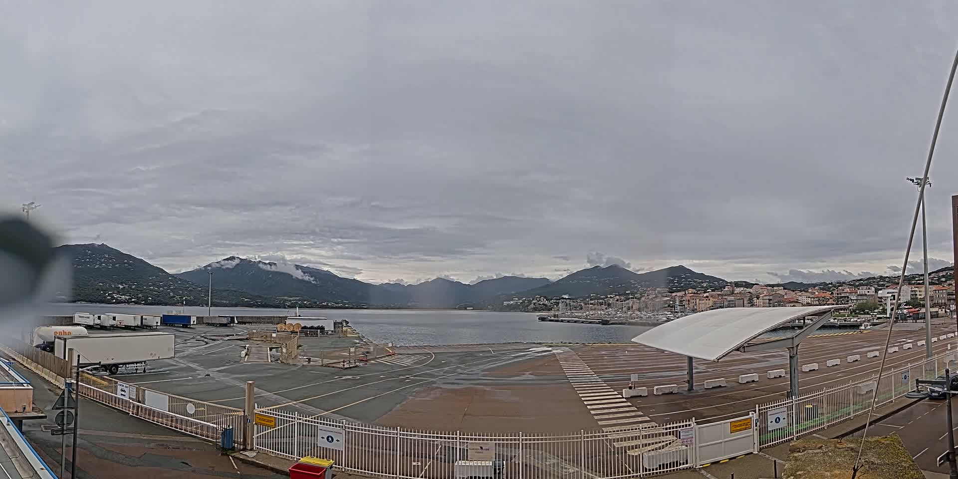 A port with several semi-trailer trucks and a covered waiting area sits in front of a calm body of water and mountains under an overcast sky.