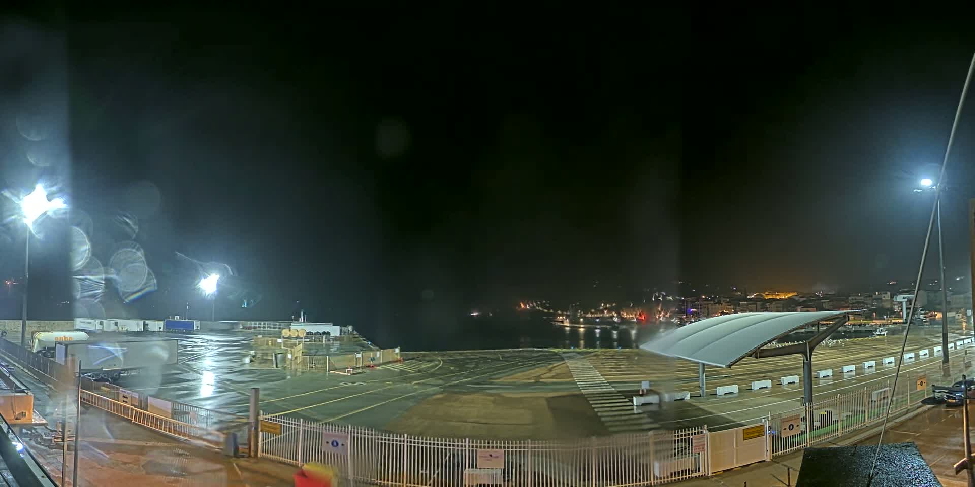 A rain-slicked, nighttime port area with bright lights illuminating a mostly empty lot, a distant town visible across the water.